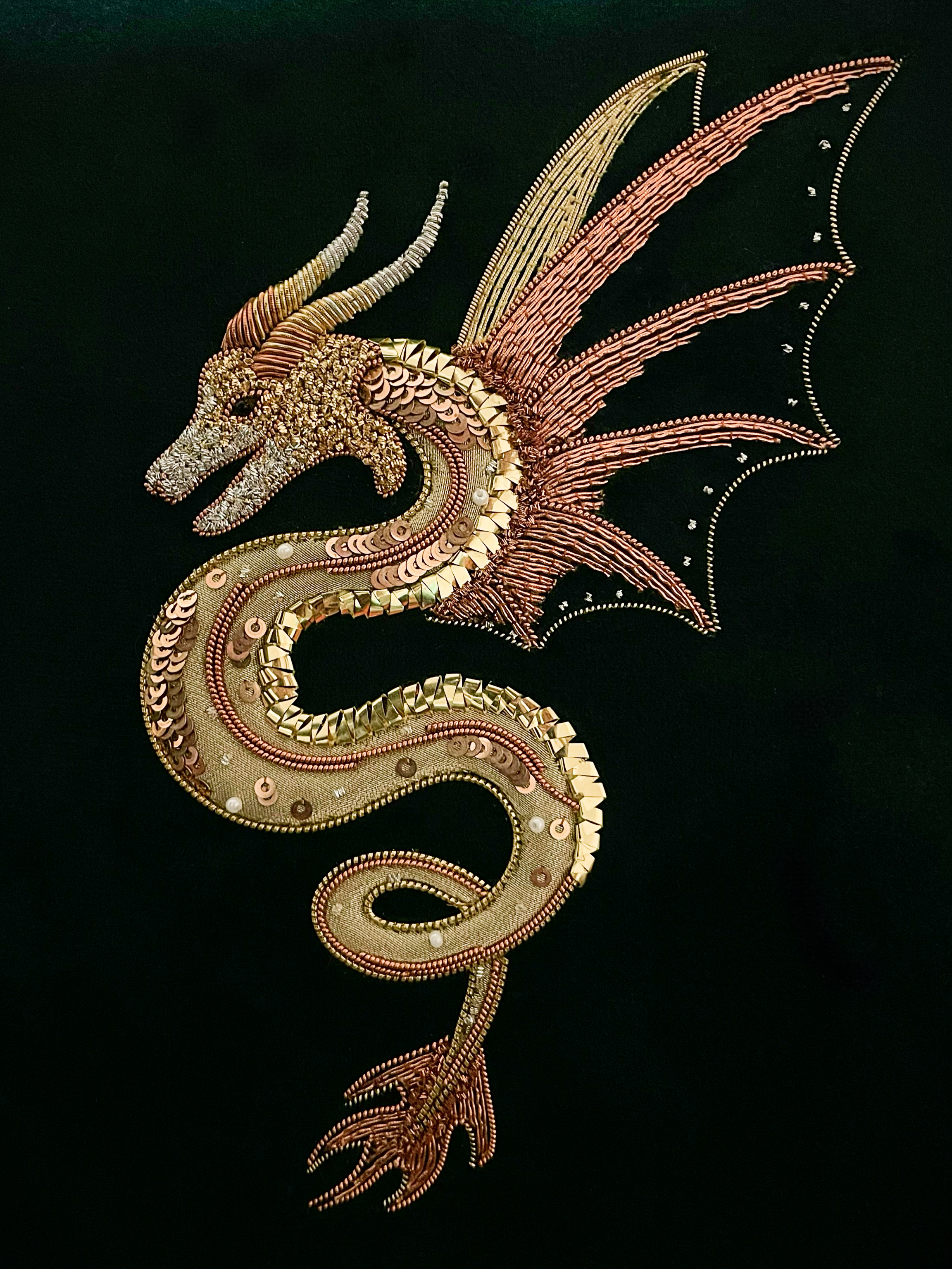 A winged dragon worked in goldwork embroidery, coiled in gold and copper metal thread with sequins and pearls on black velvet.