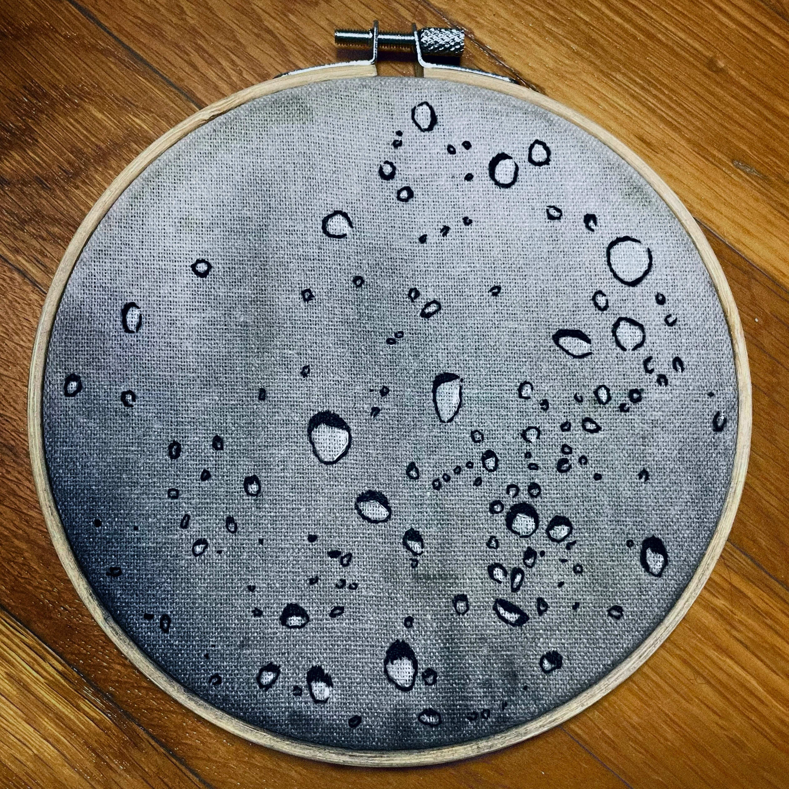 Dozens of water droplets embroidered in black thread across grey linen, scattered at different sizes so the piece reads as rain caught on a window.