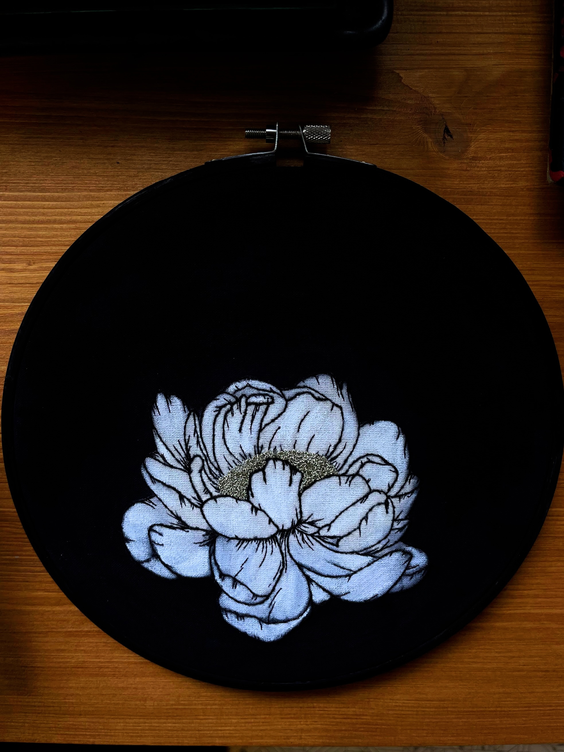 A white peony embroidered on black fabric, the petals shaded in pale thread and outlined with fine dark stitches, with a crescent of gold metal thread at the center.